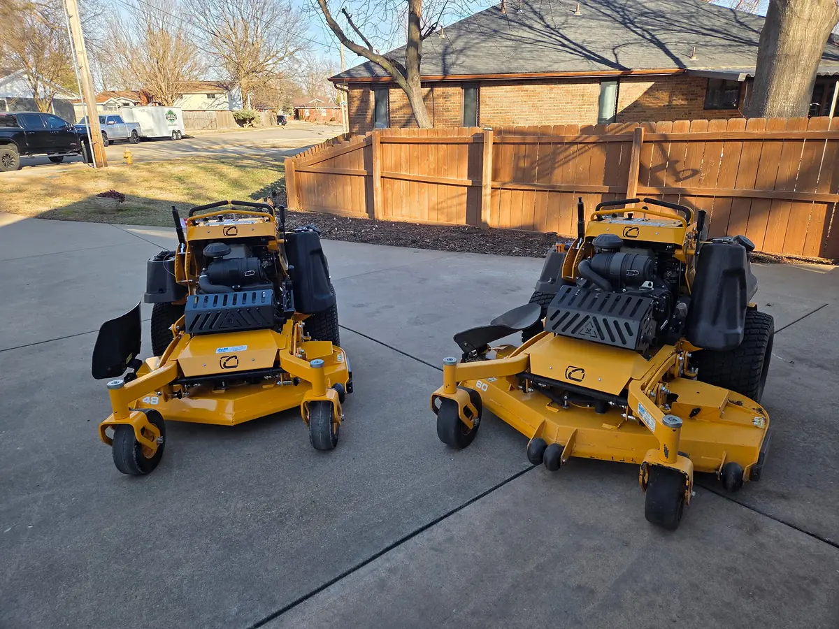 Two yellow commercial stand-on mowers on a concrete drive